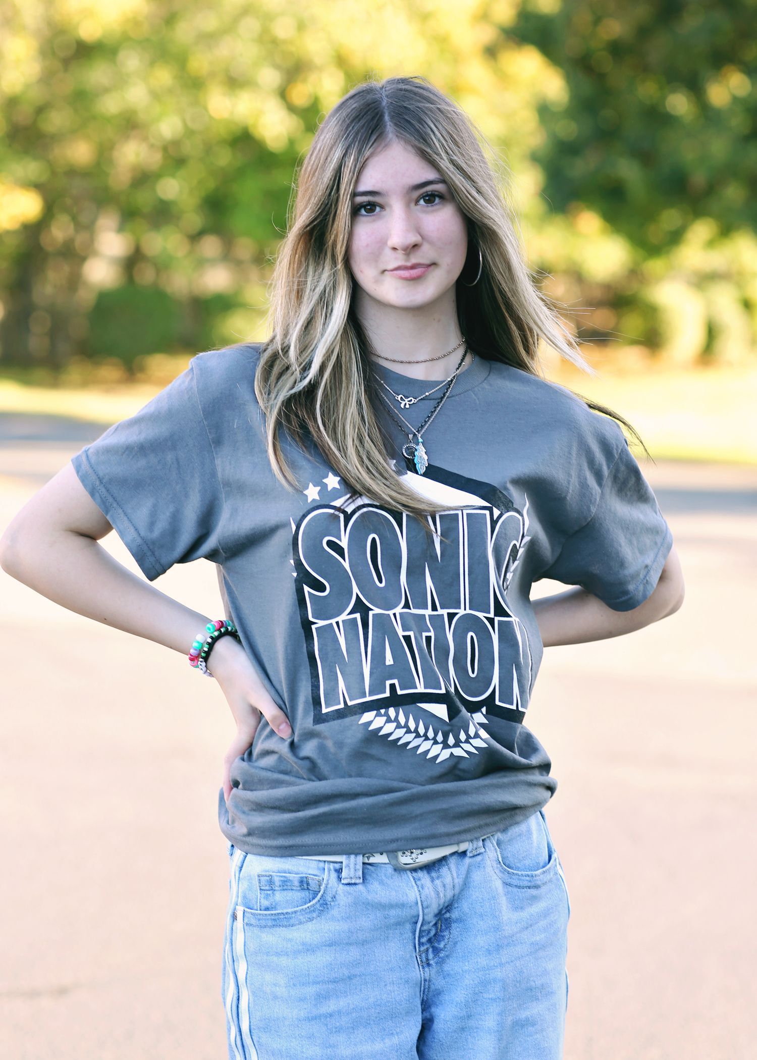 Woman with long hair in grey shirt and blue jeans, hands on hips, outdoor setting.
