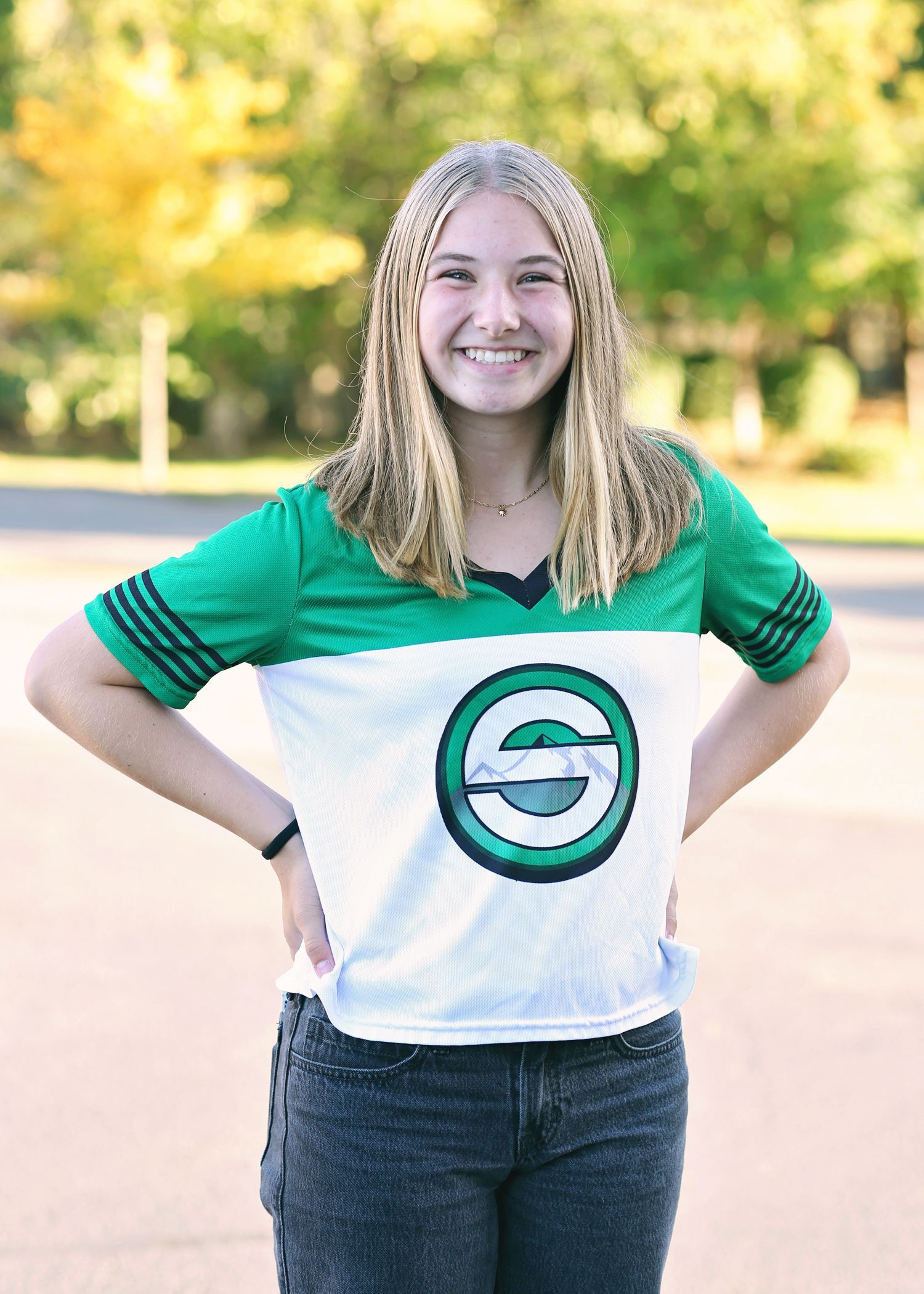 Teen girl smiling, hands on hips, wearing green and white shirt with logo, outdoors.