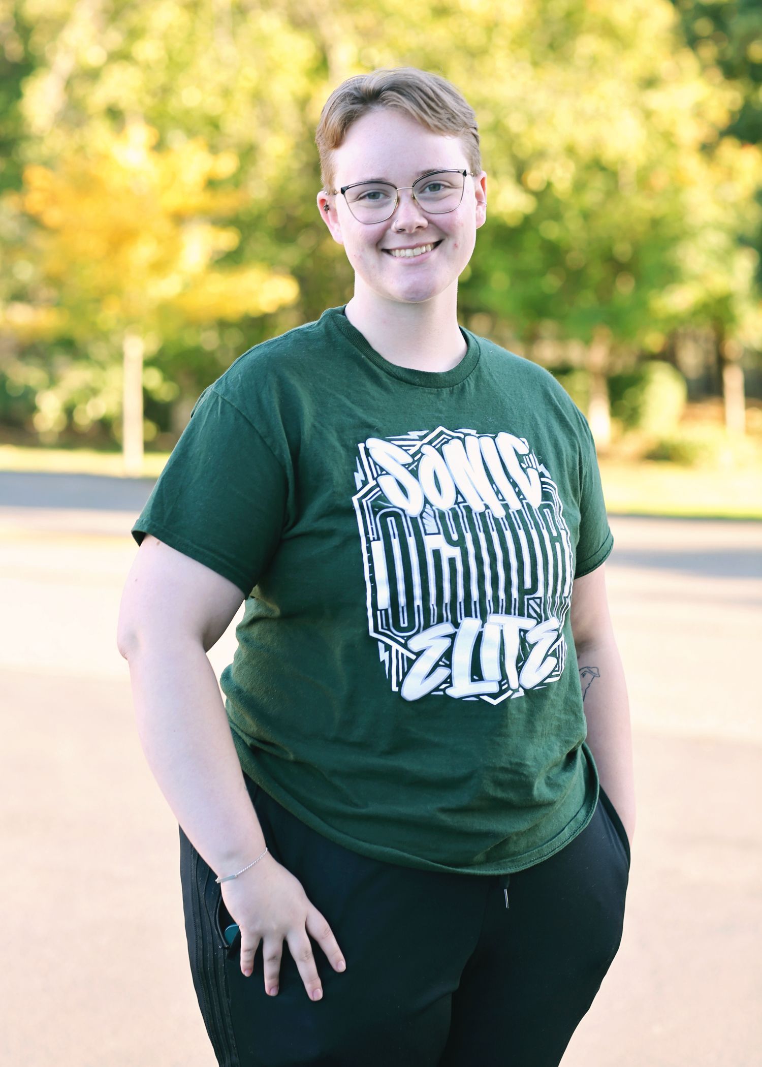 Person with short hair wearing glasses and a green shirt with a design, standing outside smiling.