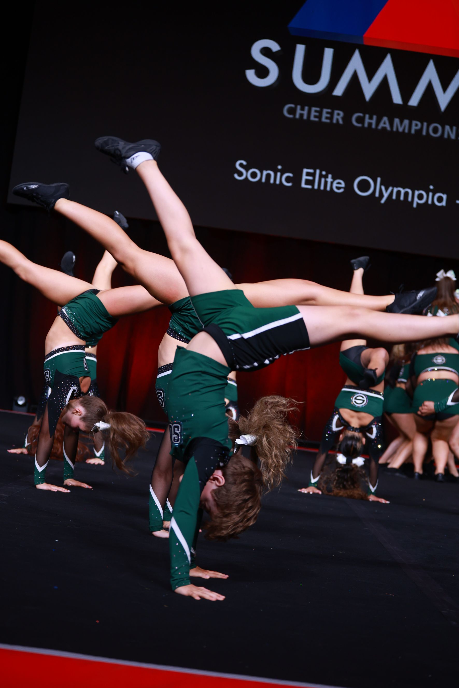 Cheerleaders doing handstands in a competition. They wear green & white uniforms and perform on a stage.
