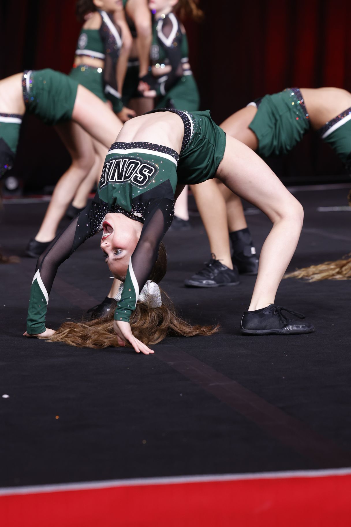 Cheerleaders in green uniforms perform a backbend on a black mat.