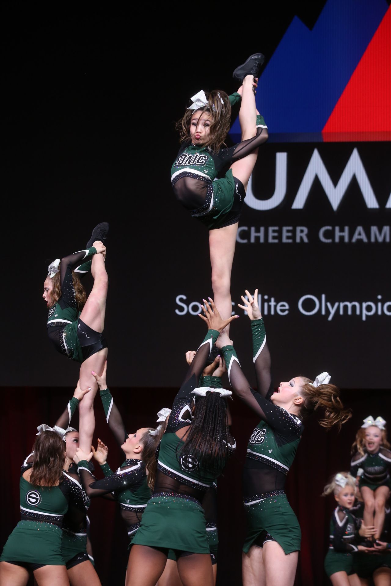 Cheerleaders performing a stunt. A flyer does a split mid-air while being supported by bases, wearing green and black uniforms at a competition.