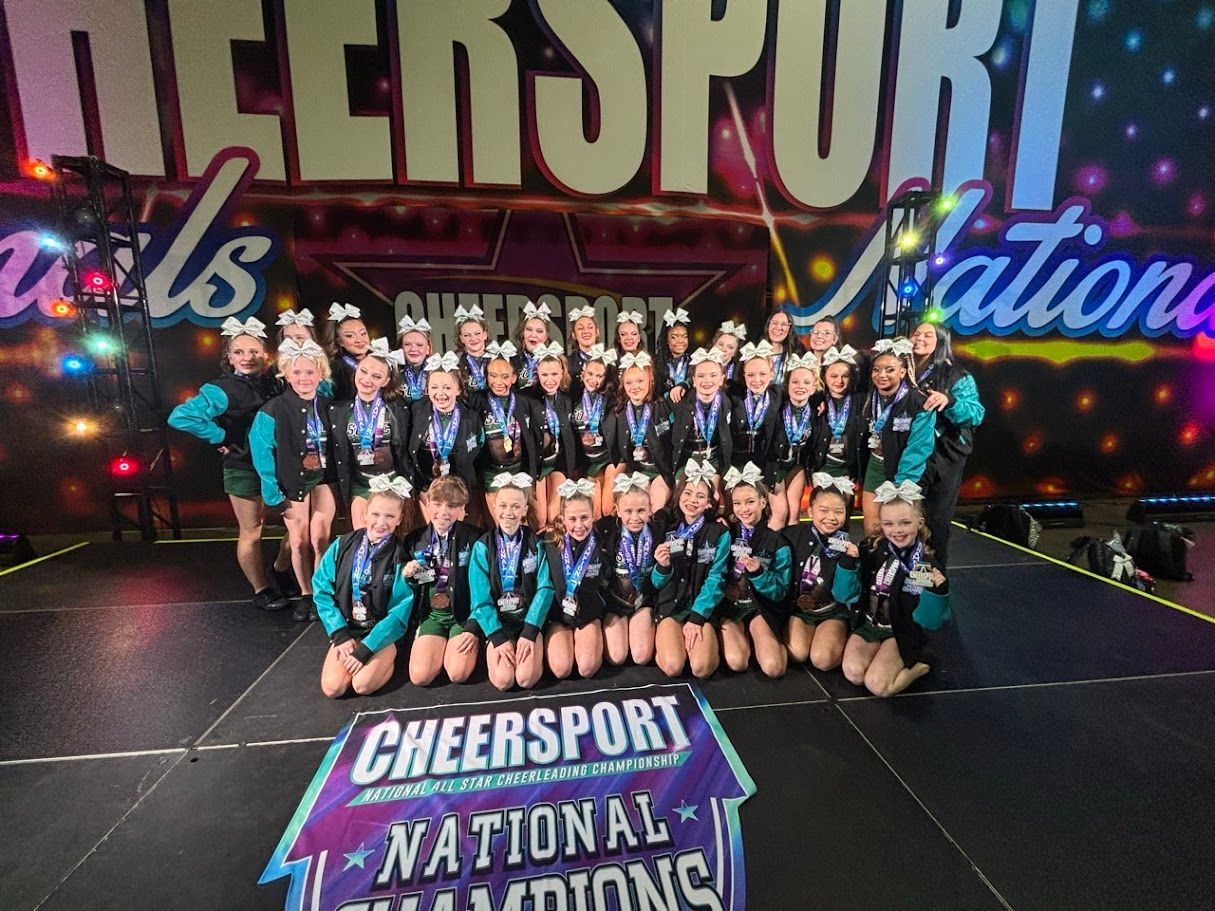 Cheer team posing on stage after winning at Cheersport Nationals. Turquoise and black uniforms, medals, and smiles.