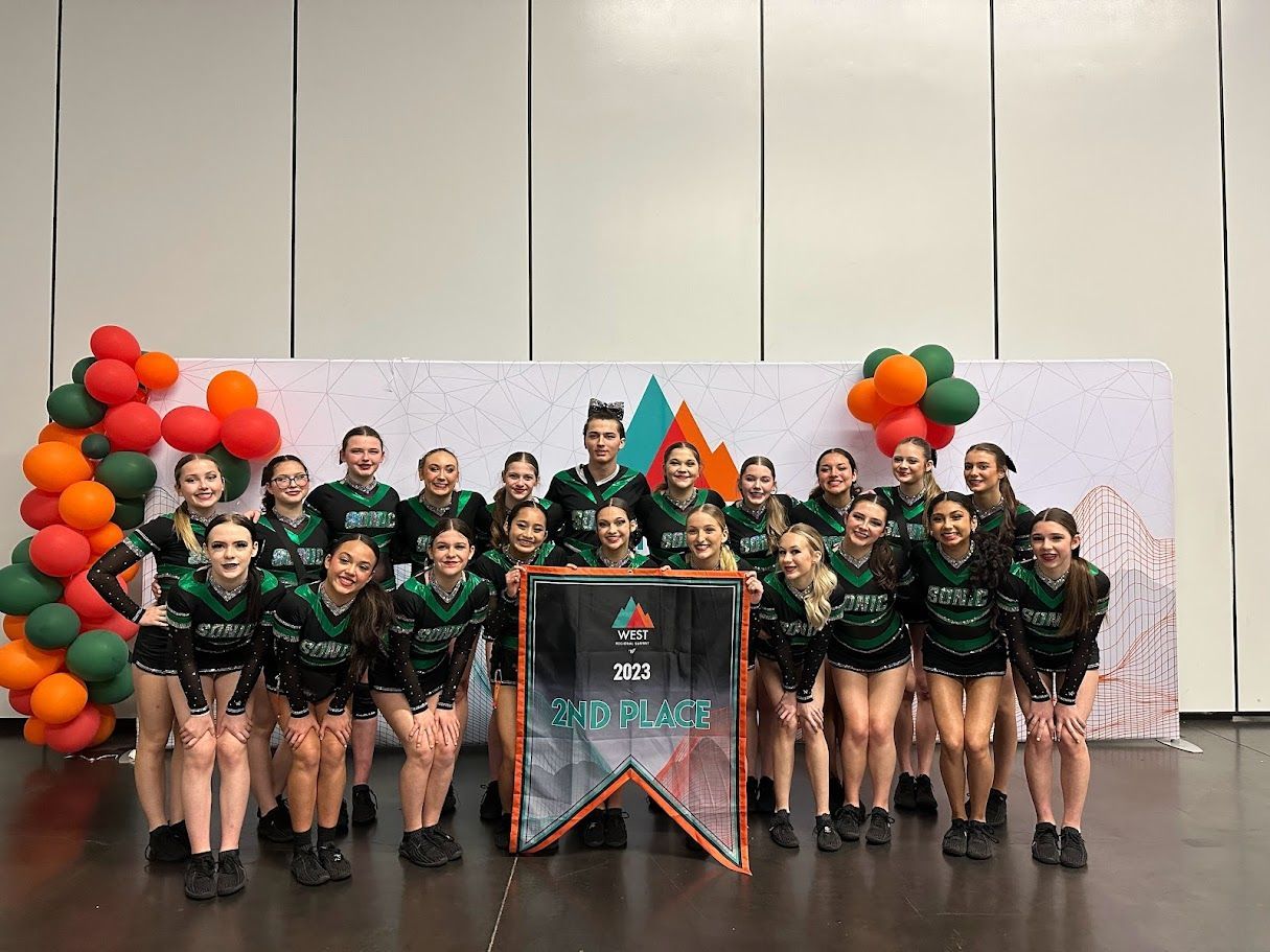 Cheerleading team poses with banner. They wear green and black uniforms and stand in front of a backdrop with orange/green balloons.