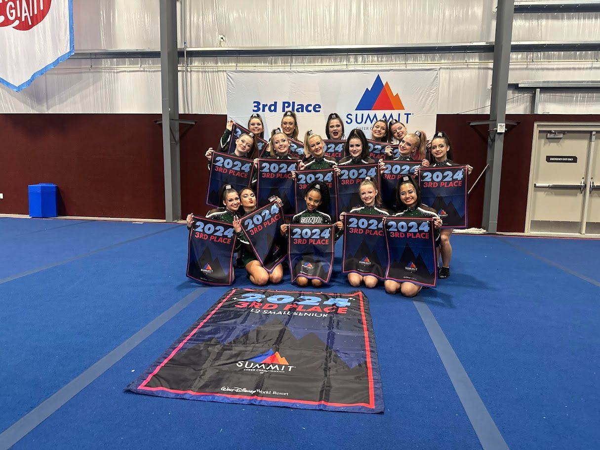 Cheerleading team posing with 3rd Place Summit banner, holding signs. Wearing black and blue uniforms, smiling in a gym.