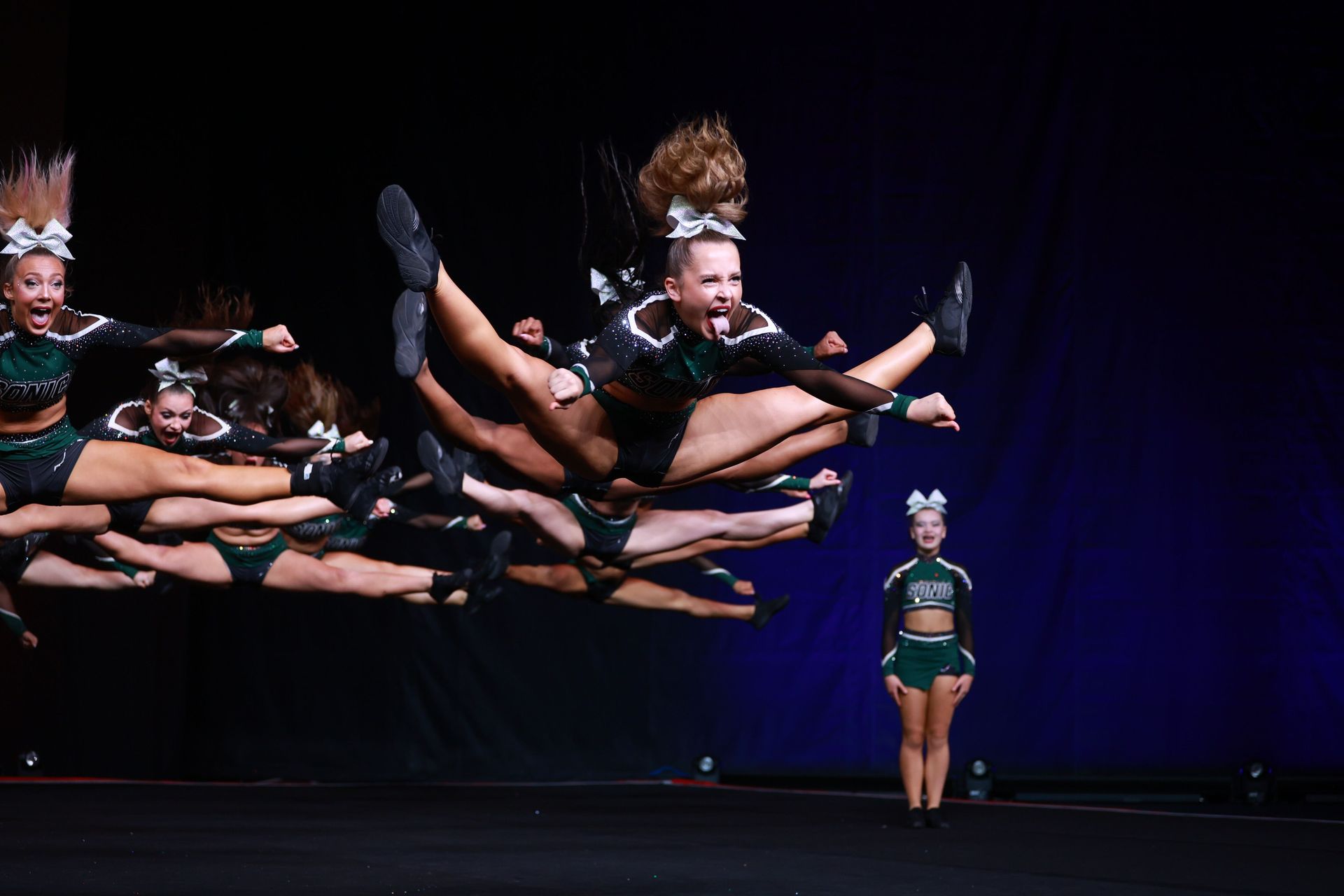 Cheerleaders in green uniforms jumping mid-air. A girl at the front has a wild hair, smiling. Black background.