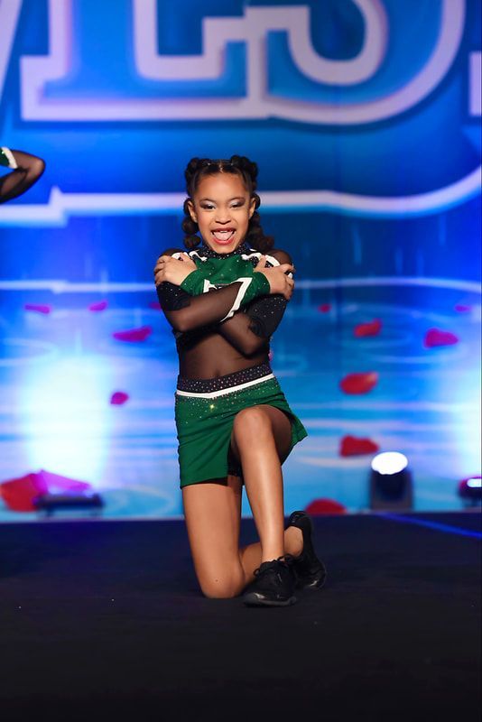 A young cheerleader kneels, arms crossed, smiling. Wearing green and black uniform on a stage.