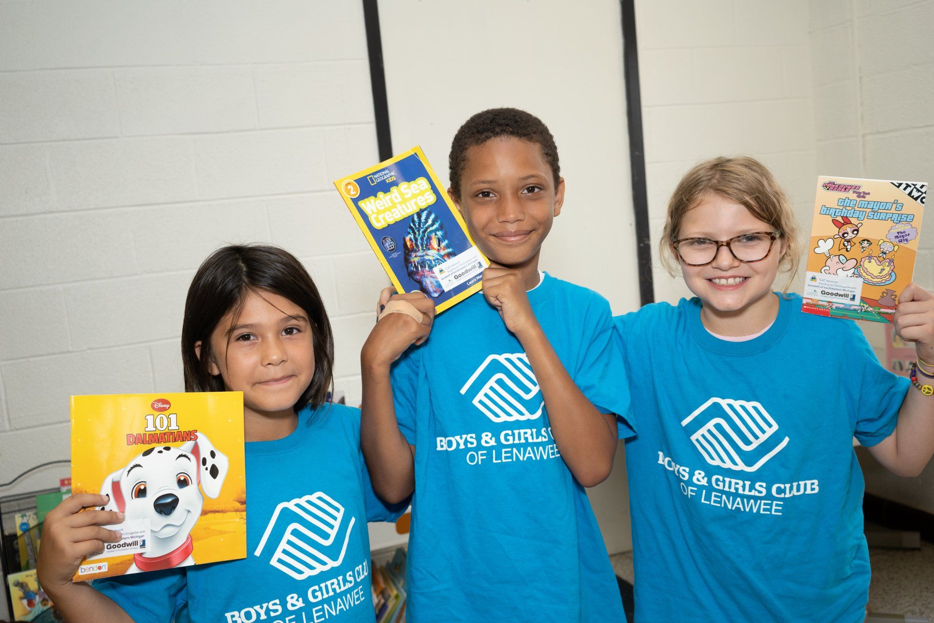 Three kids holding books wearing blue Boys & Girls Club of Lenawee shirts