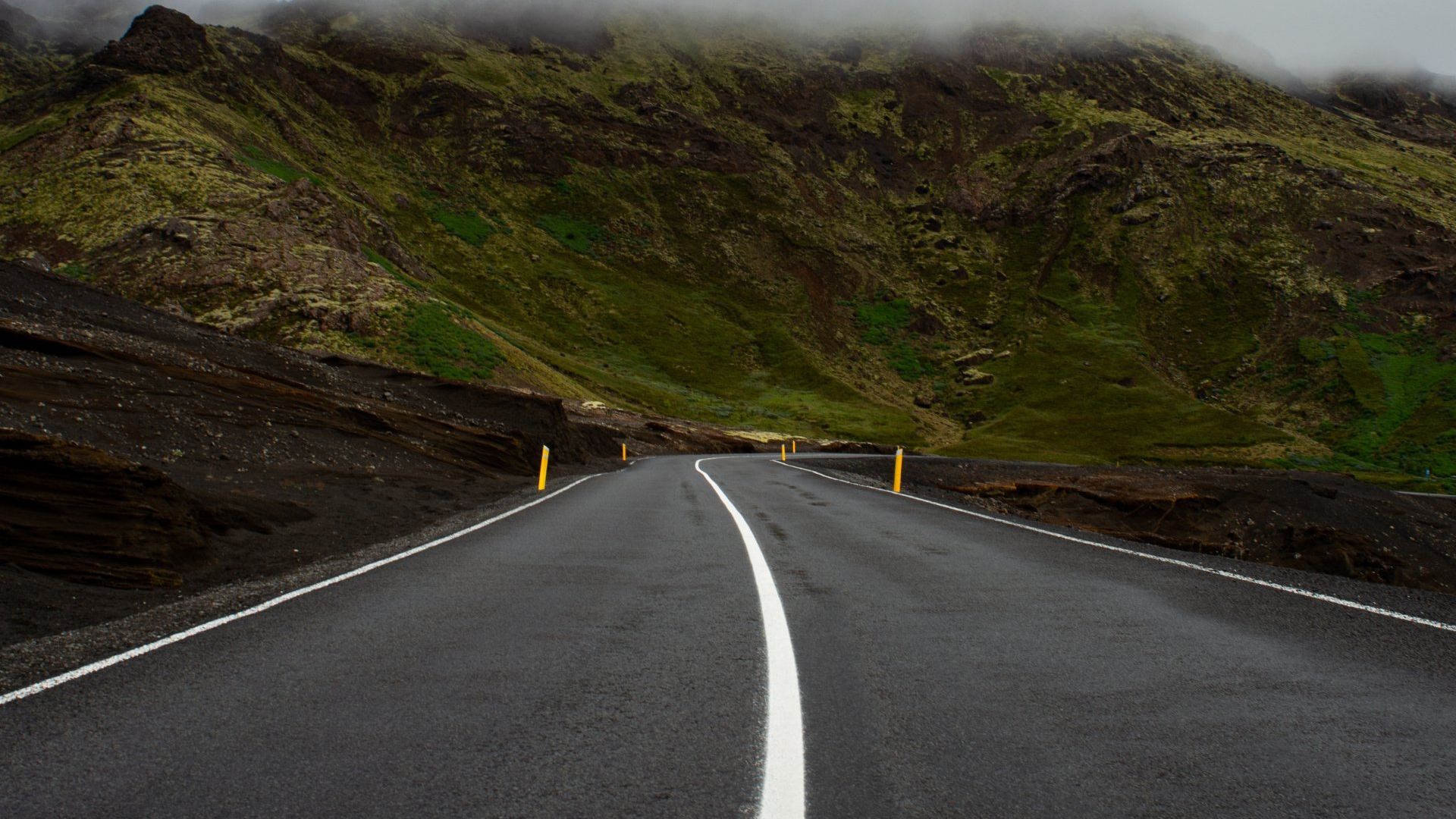 A paved road with a white center line winds through a rugged landscape of dark volcanic rock and green, misty mountains.