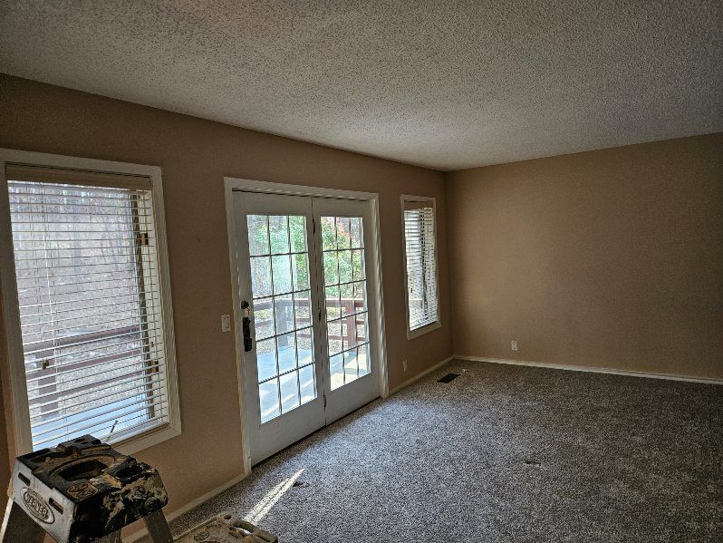 Empty room with beige walls, gray carpet, and a pair of French doors, and two windows with blinds.