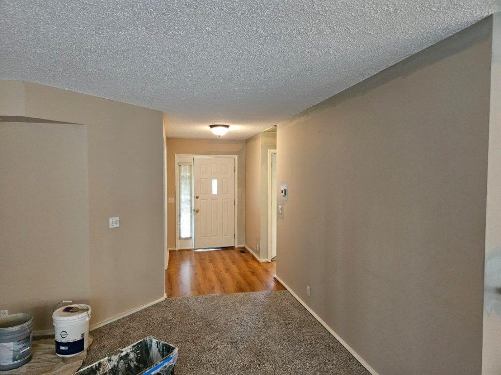 Beige-painted interior hallway with entry door. Tan walls, carpet, popcorn ceiling; paint supplies in foreground.