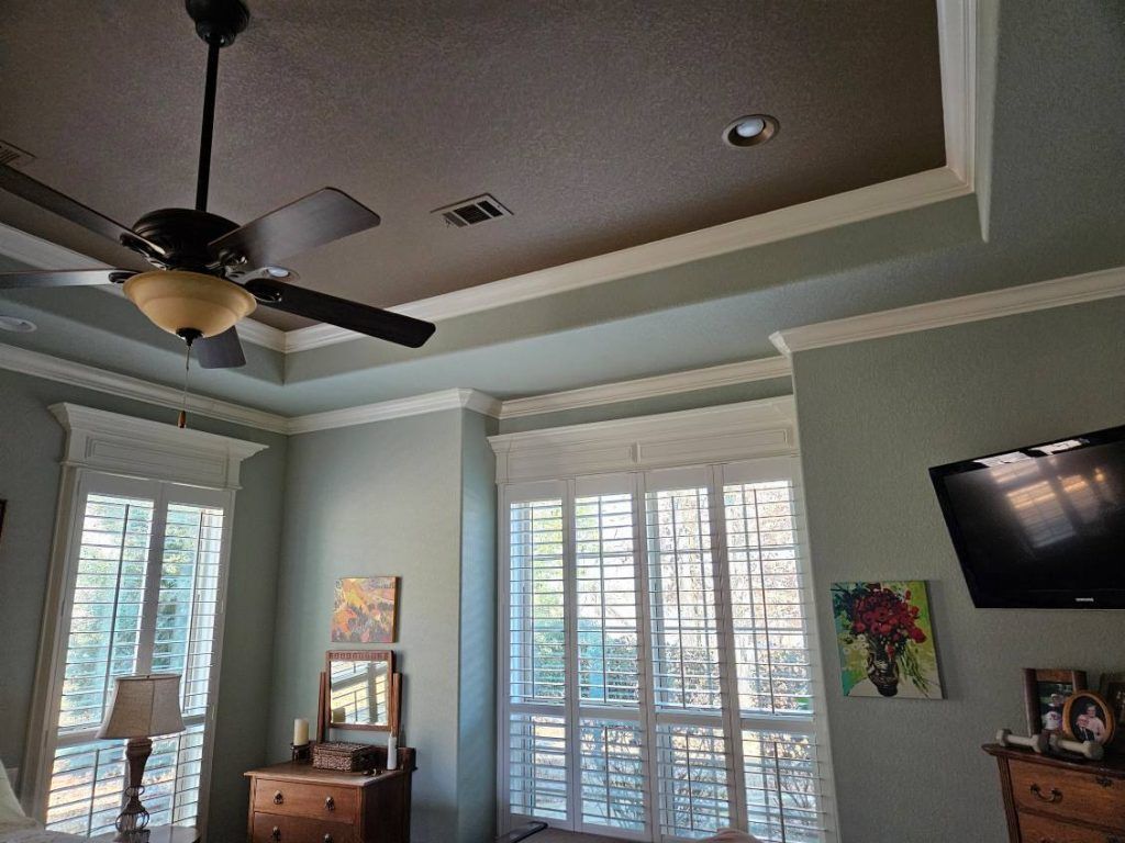 A bedroom with blue walls, a ceiling fan, windows with white shutters, and a mounted TV.