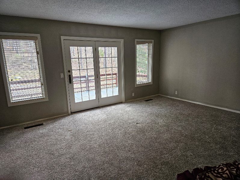 Empty room with gray walls, carpet, and white door and window frames overlooking a deck and trees.