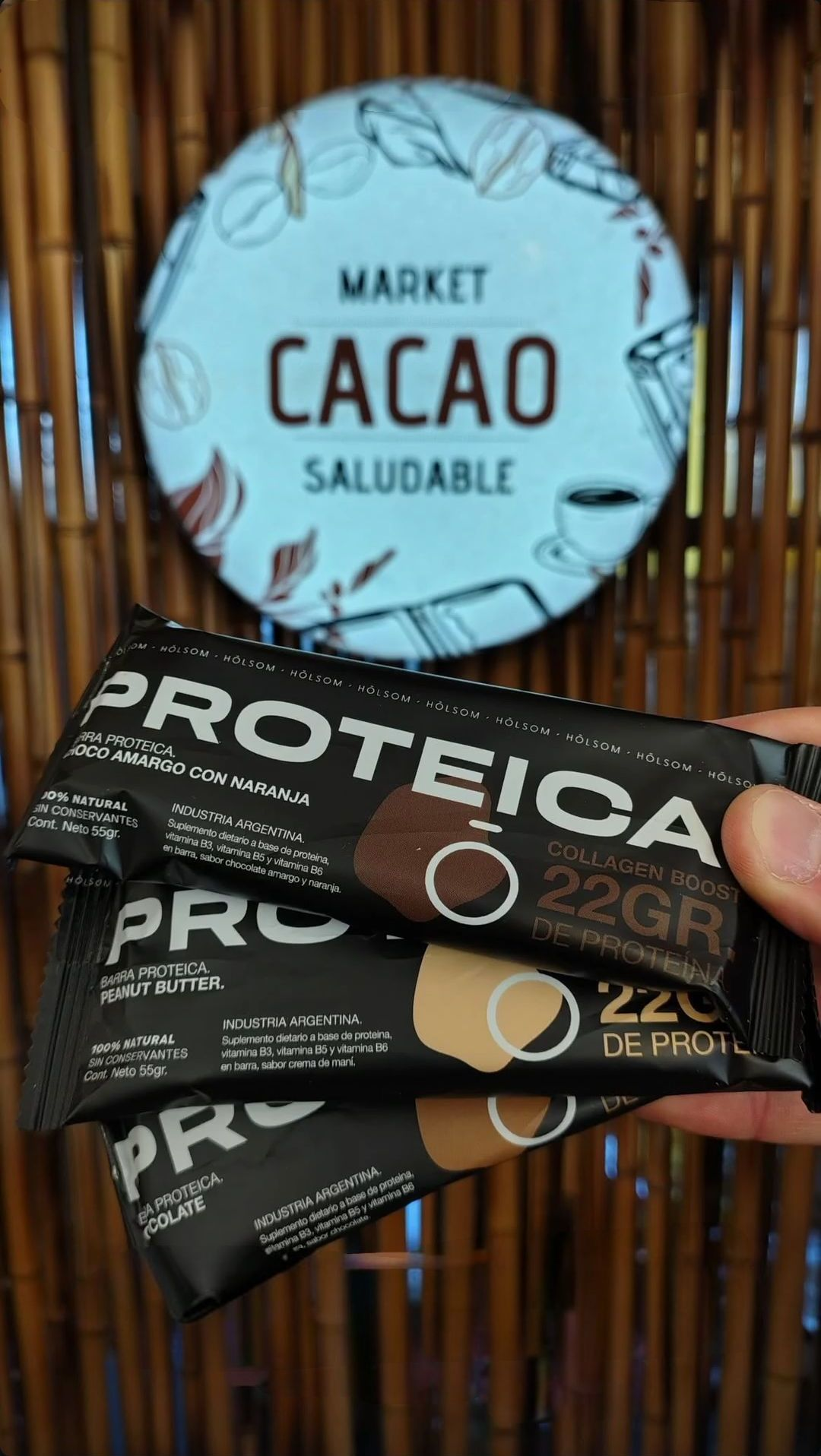 Mano sosteniendo tres barras de proteína oscuras frente a un letrero de “Mercado de Cacao”.
