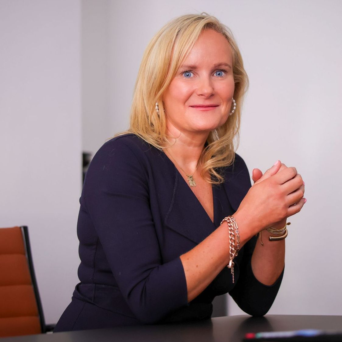 A woman in a blue dress sits at a table with her hands folded