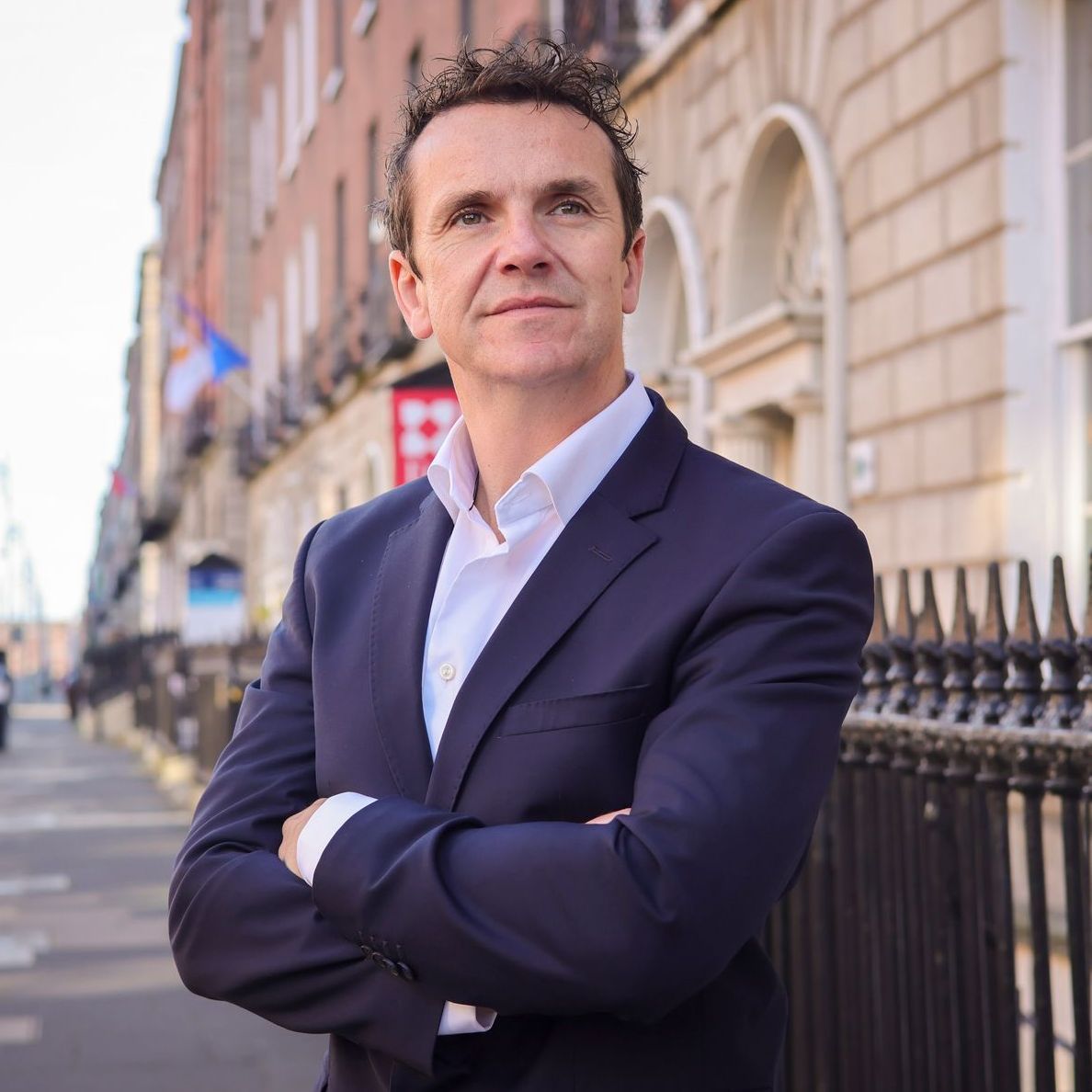 A man in a suit is standing with his arms crossed in front of a building.