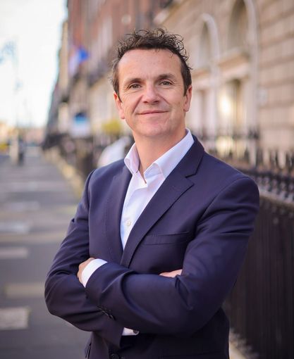 A man in a suit and white shirt is standing with his arms crossed in front of a building.