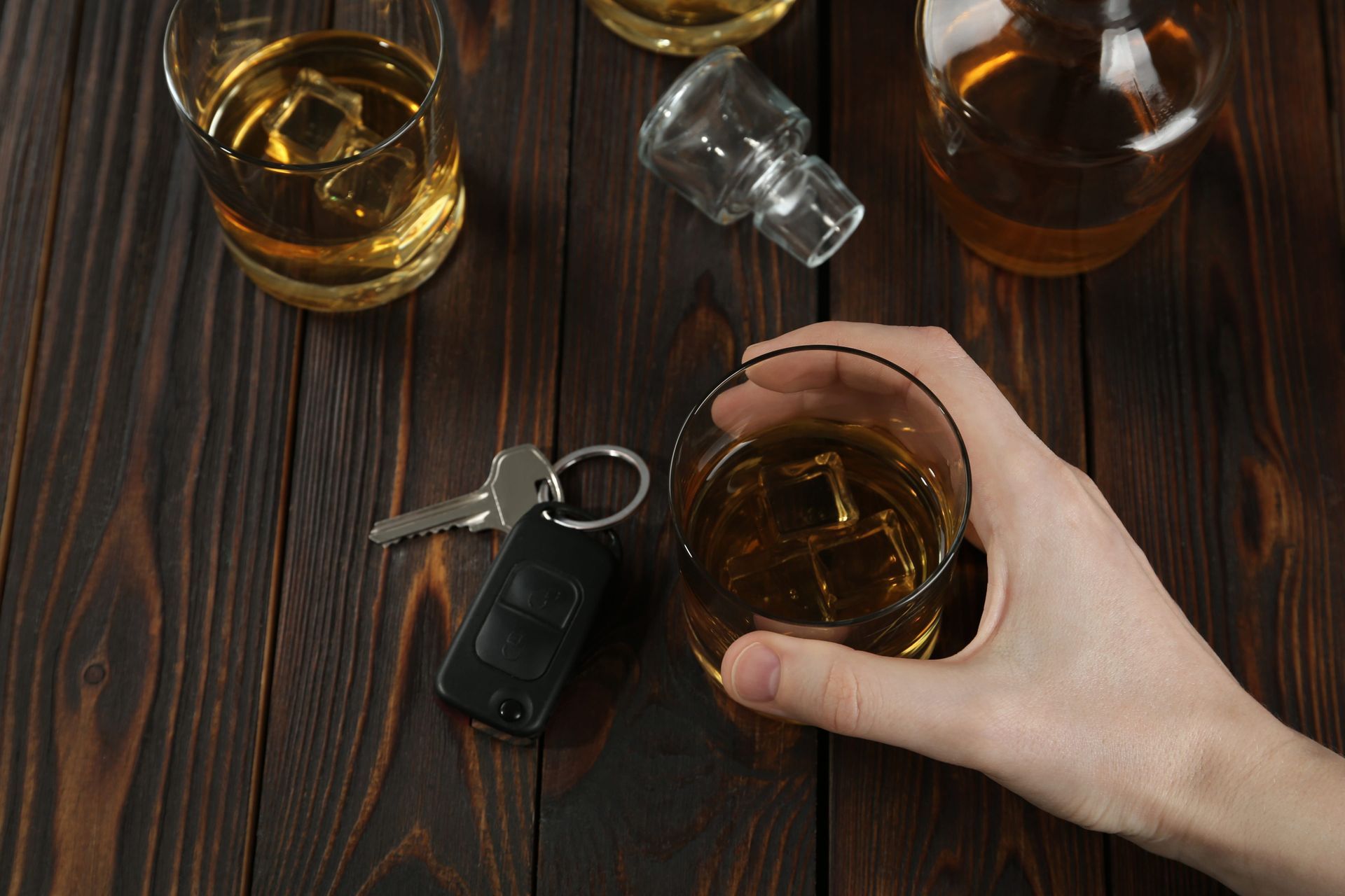 A hand holds a glass of whiskey next to car keys and a bottle on a wooden table, suggesting drunk driving.