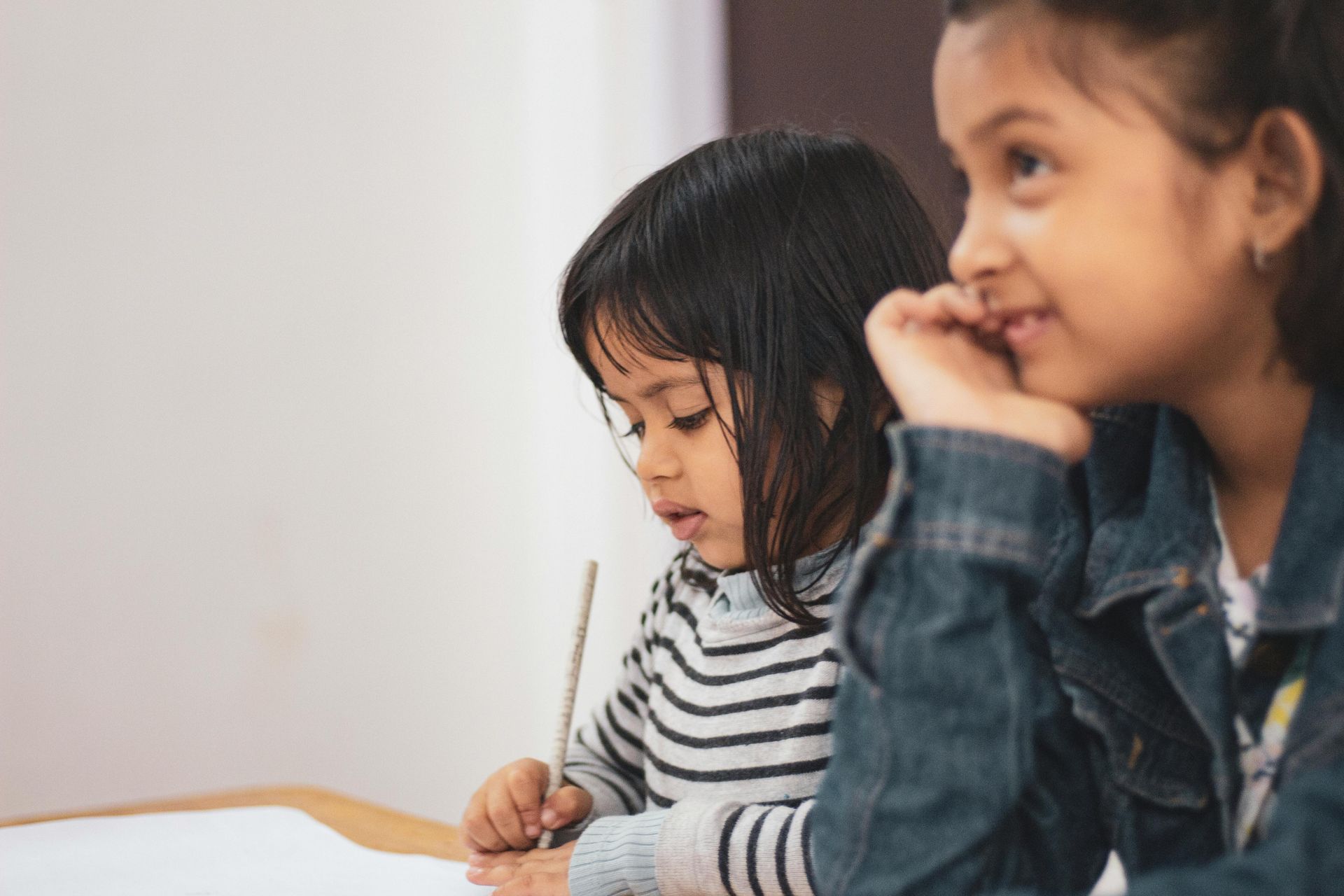 Two little girls are sitting at a table writing on a piece of paper.