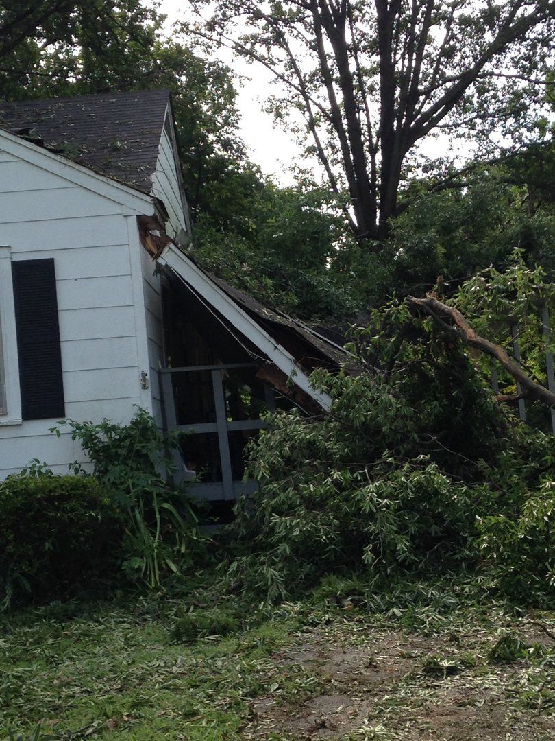 A white house with a damaged porch covered in fallen tree branches and foliage.