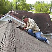 Man on roof inspecting shingles, wearing jeans, tan jacket, and cap.