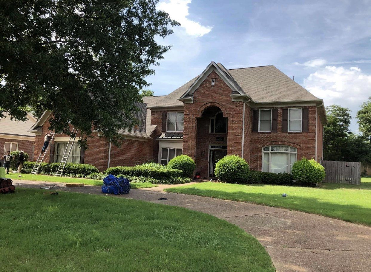 Red brick house with a gray roof and green lawn. Two people on a ladder.