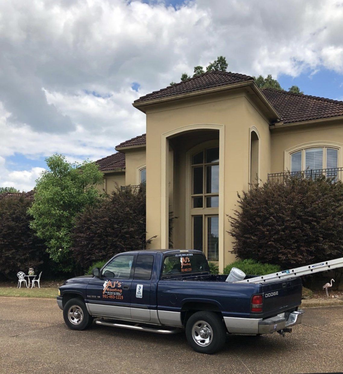 Dark blue pickup truck parked in front of a large beige house, with a ladder in the truck bed.
