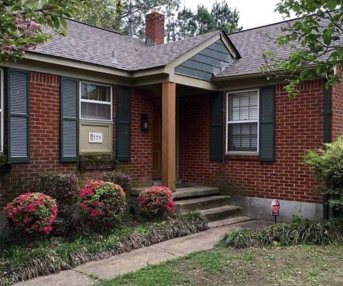 Red brick house with green shutters, steps, and flowering bushes in front.