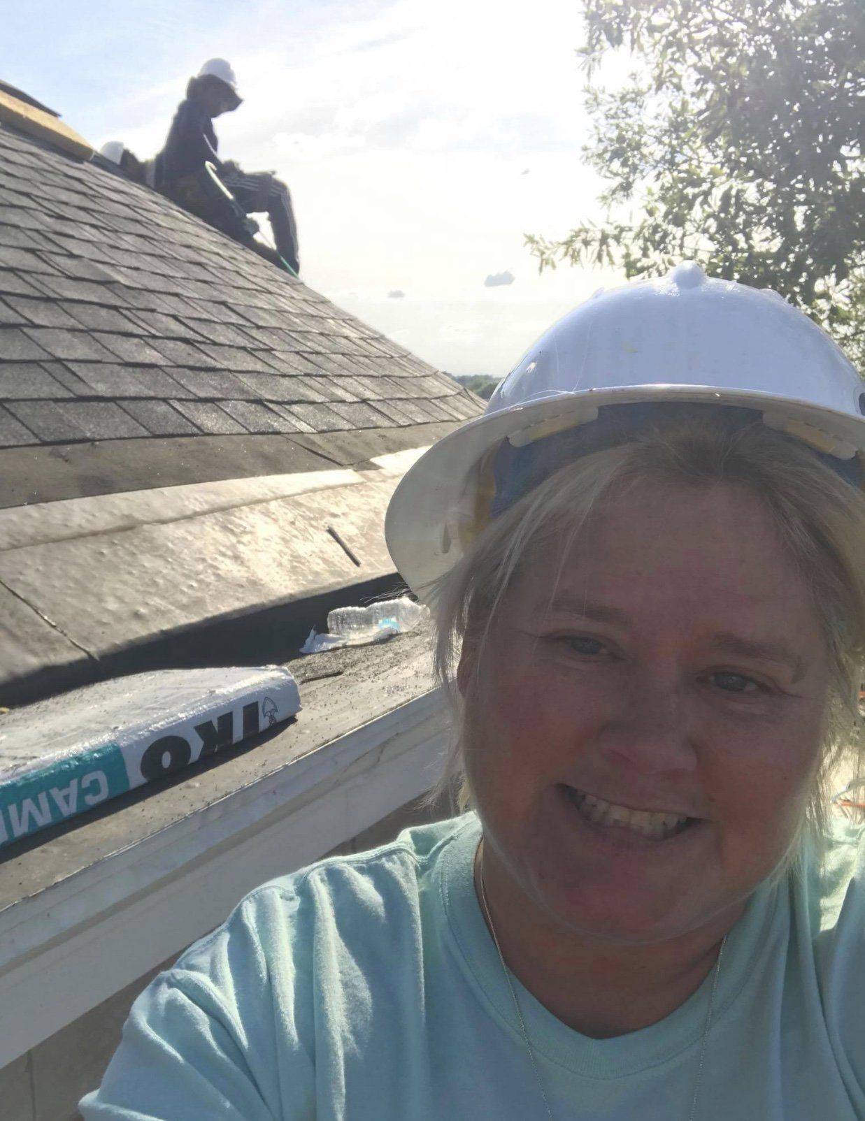Woman in hard hat smiles for selfie on a roof as roofers work in the background.