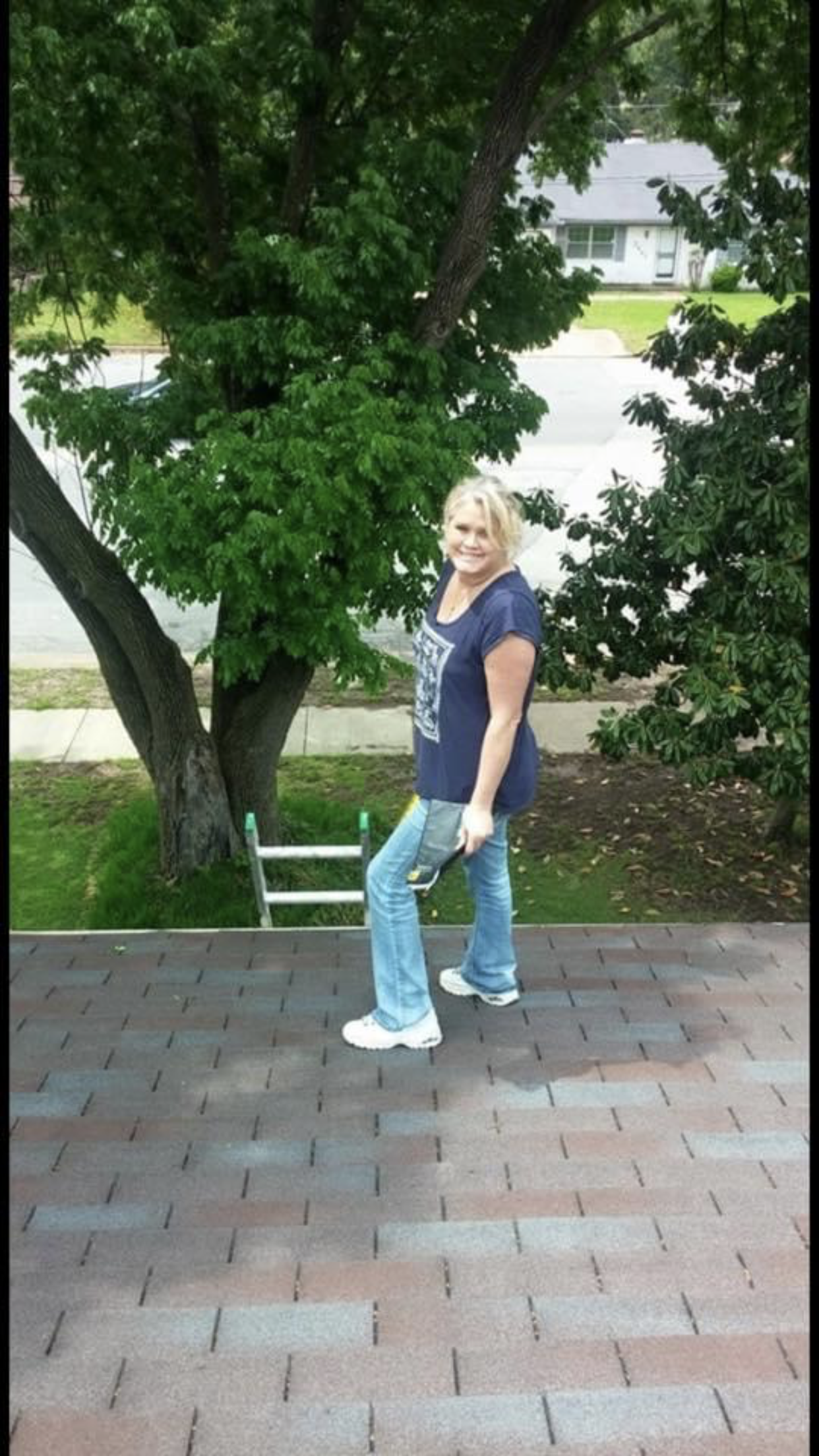 Woman on a roof with a tool, wearing blue jeans and a t-shirt, near a tree in a residential area.