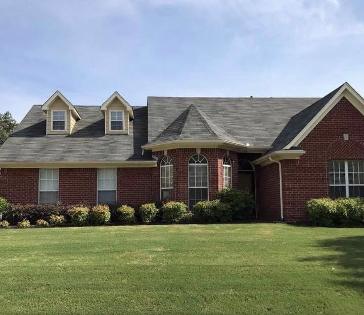 Brick house with green lawn, bushes, and a gray roof under a blue sky.