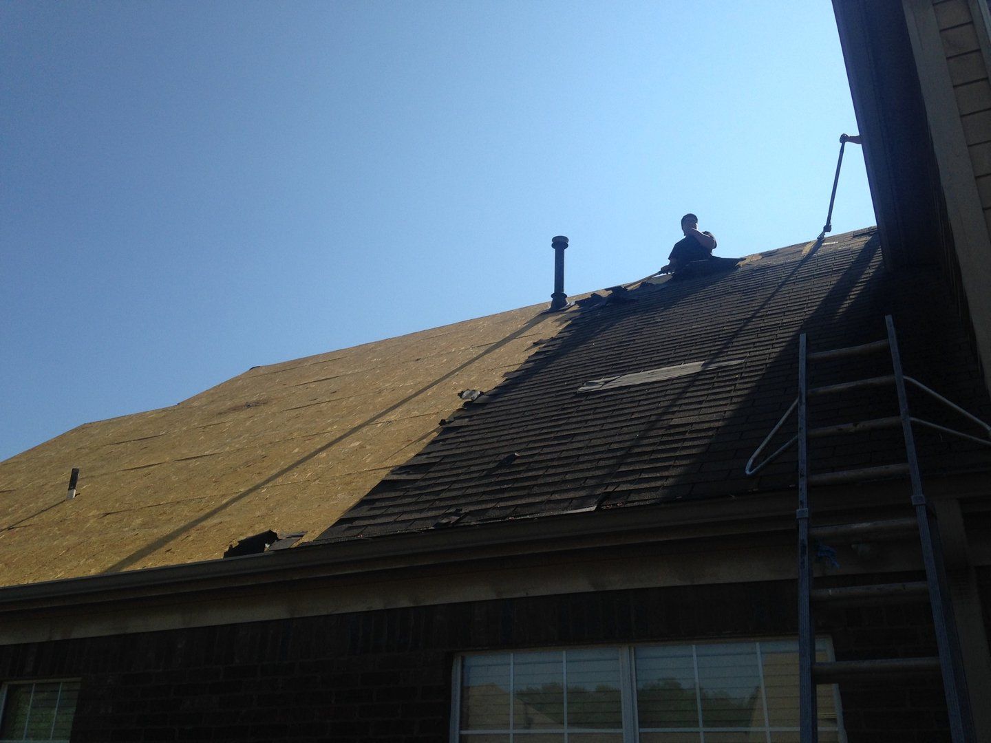 Person on a roof replacing shingles. Partial roof replacement in progress. Ladder visible. Blue sky.