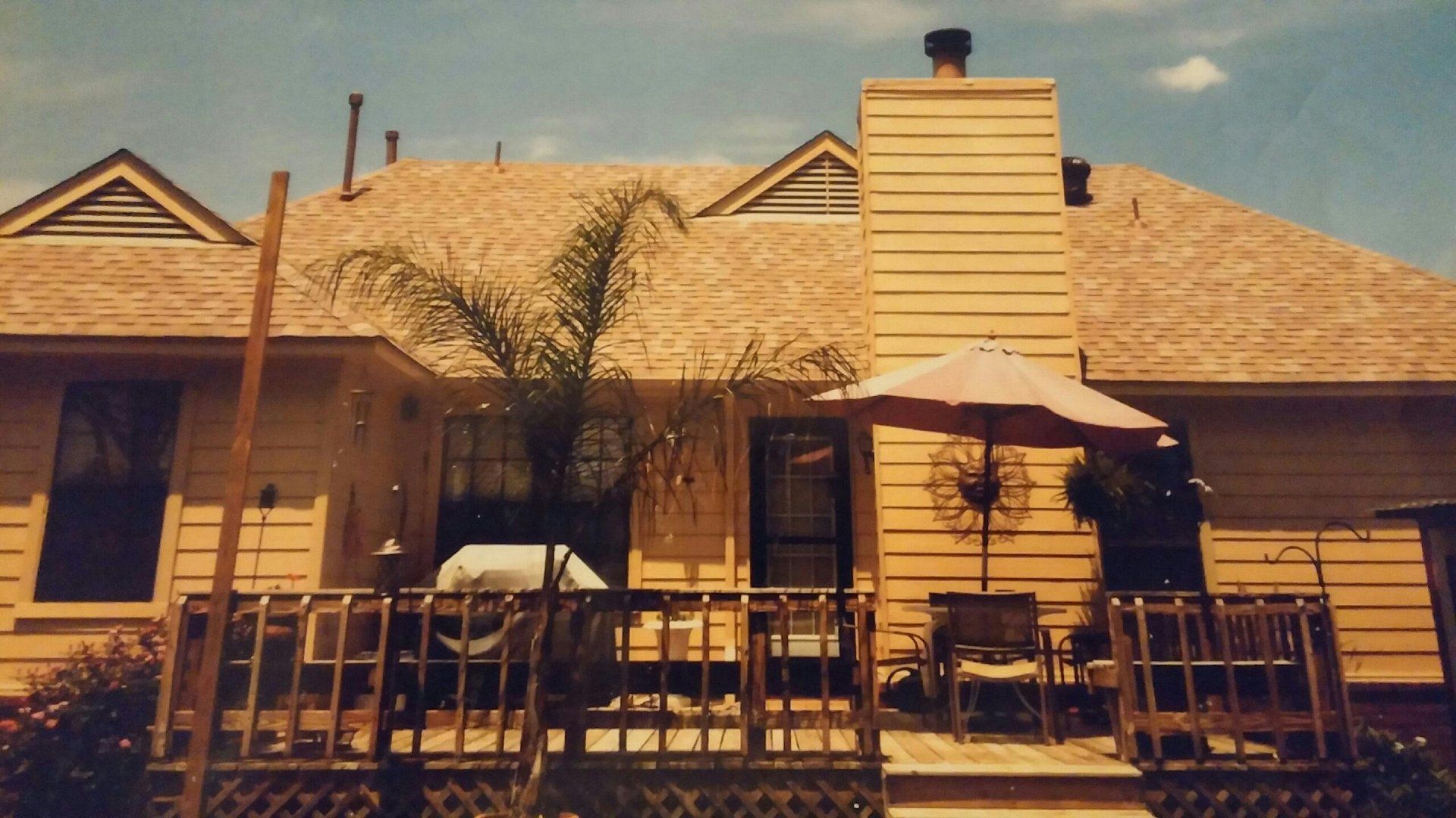 Backyard deck of a yellow house with a palm tree, umbrella, and chimney against a blue sky.