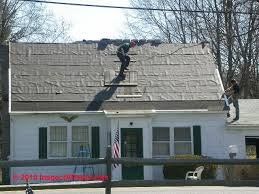 Two workers on a roof, removing shingles. White house with green shutters, an American flag, and trees.