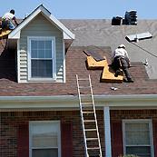 Two roofers replacing shingles on a house with a dormer. A ladder is leaning against the roof.