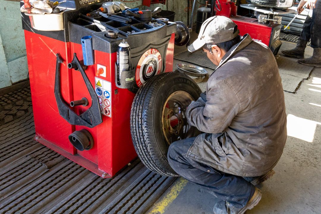 Mechanic balances a tire on a red machine in a garage. He's wearing a cap and overalls.