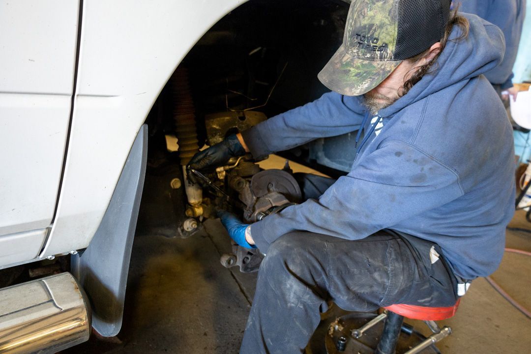 Mechanic working on a vehicle's wheel well, wearing gloves, camo hat, blue hoodie.