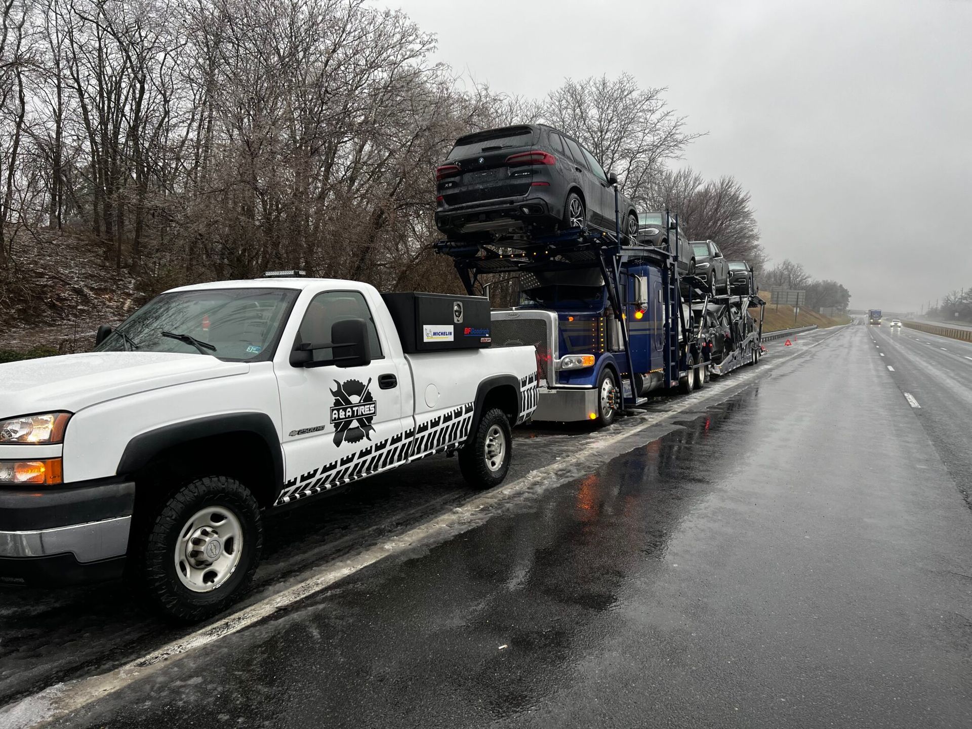 White pickup truck pulling a car carrier loaded with vehicles on a wet highway.