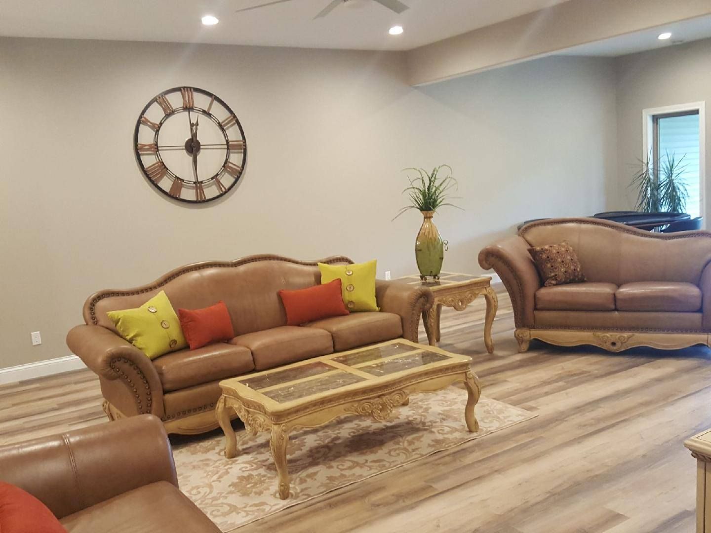 Living room with brown sofas, yellow and orange pillows, glass coffee table, wall clock, and wood flooring