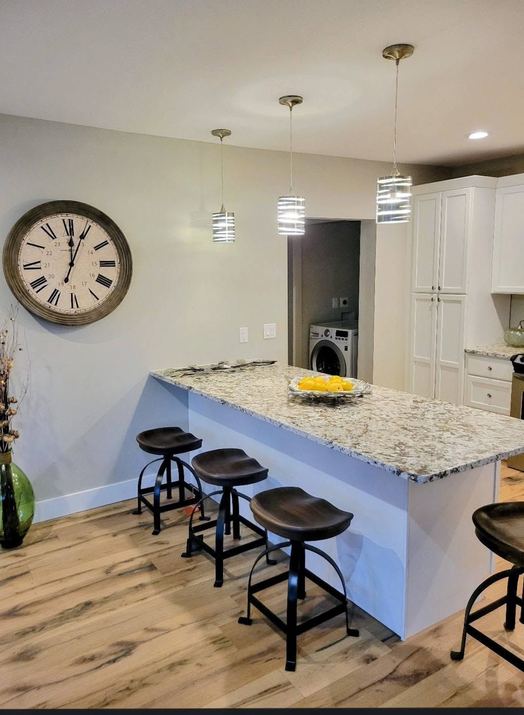Modern kitchen with granite island, three stools, pendant lights, and large wall clock on the left.