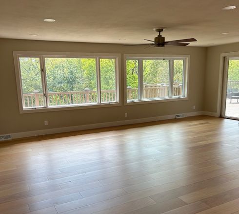Empty living room with hardwood floors, large windows, and a ceiling fan, filled with natural light
