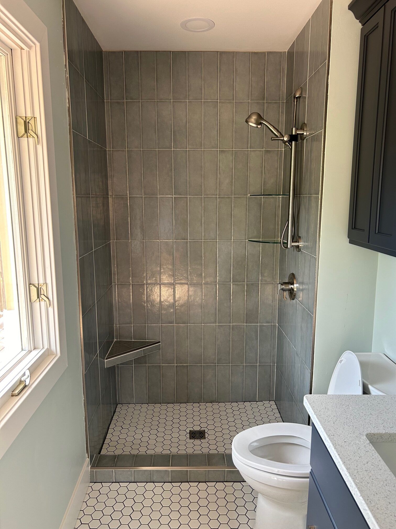 Small tiled shower with grab bars and shower chair beside a toilet in a bathroom