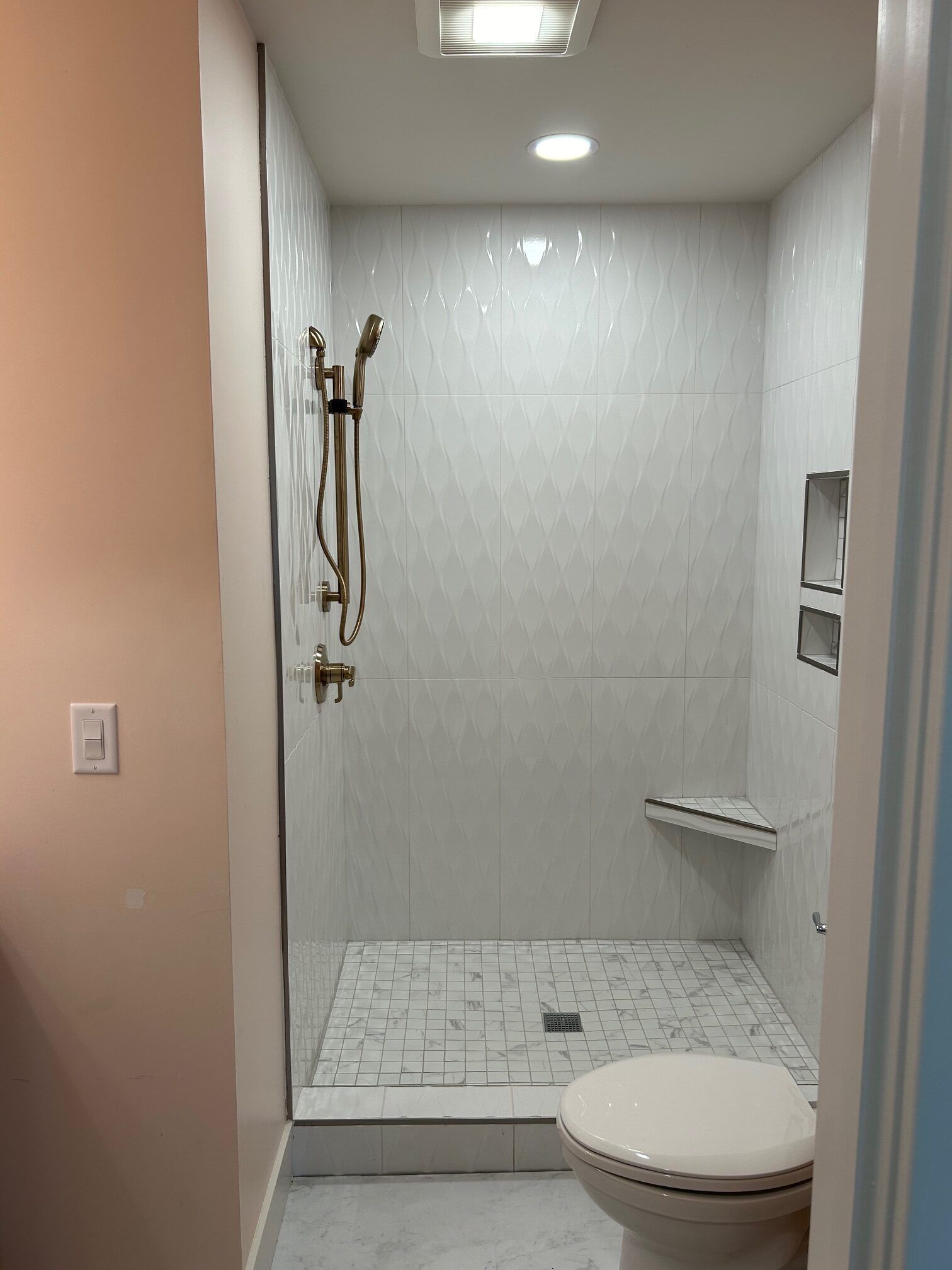 Small white tiled shower stall with grab bars and built-in seat beside a toilet in a bathroom