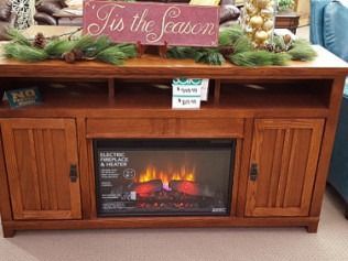 Wooden electric fireplace display with lit flames and a “Tis the Season” holiday sign on top