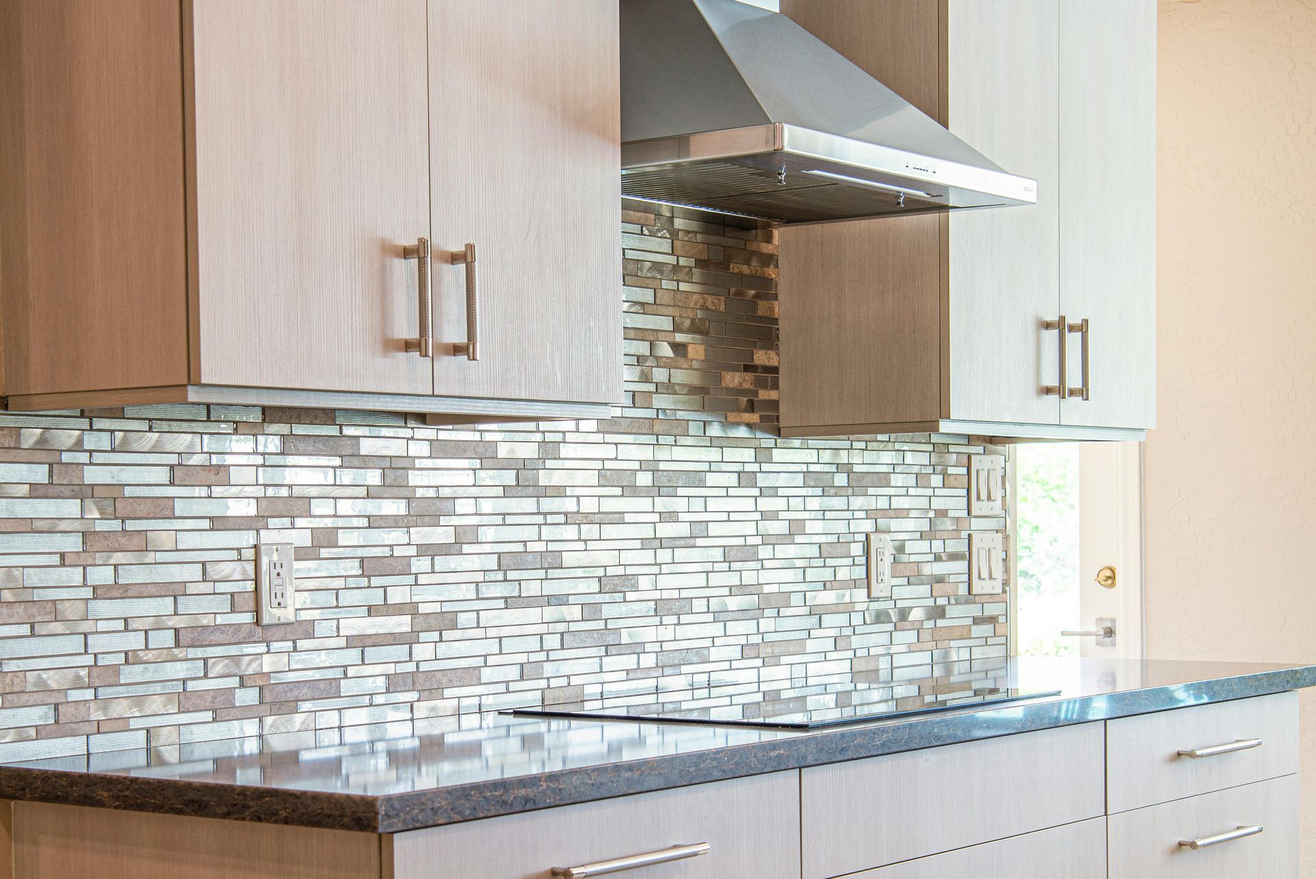 Modern kitchen with wood cabinets, mosaic tile backsplash, stainless range hood, and black countertop