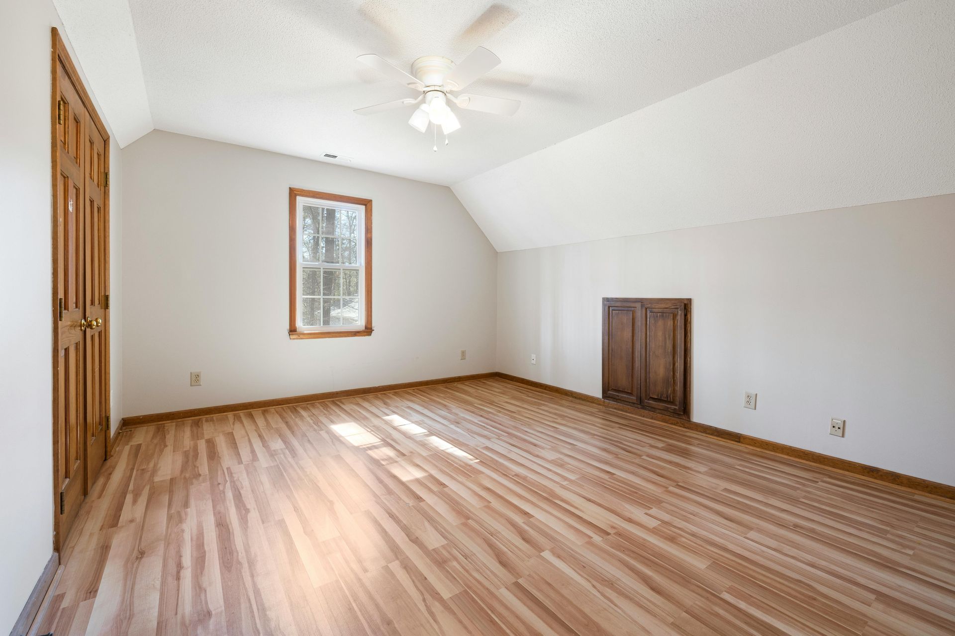 Empty carpeted bedroom with white walls, wood floor, ceiling fan, and a small framed window.