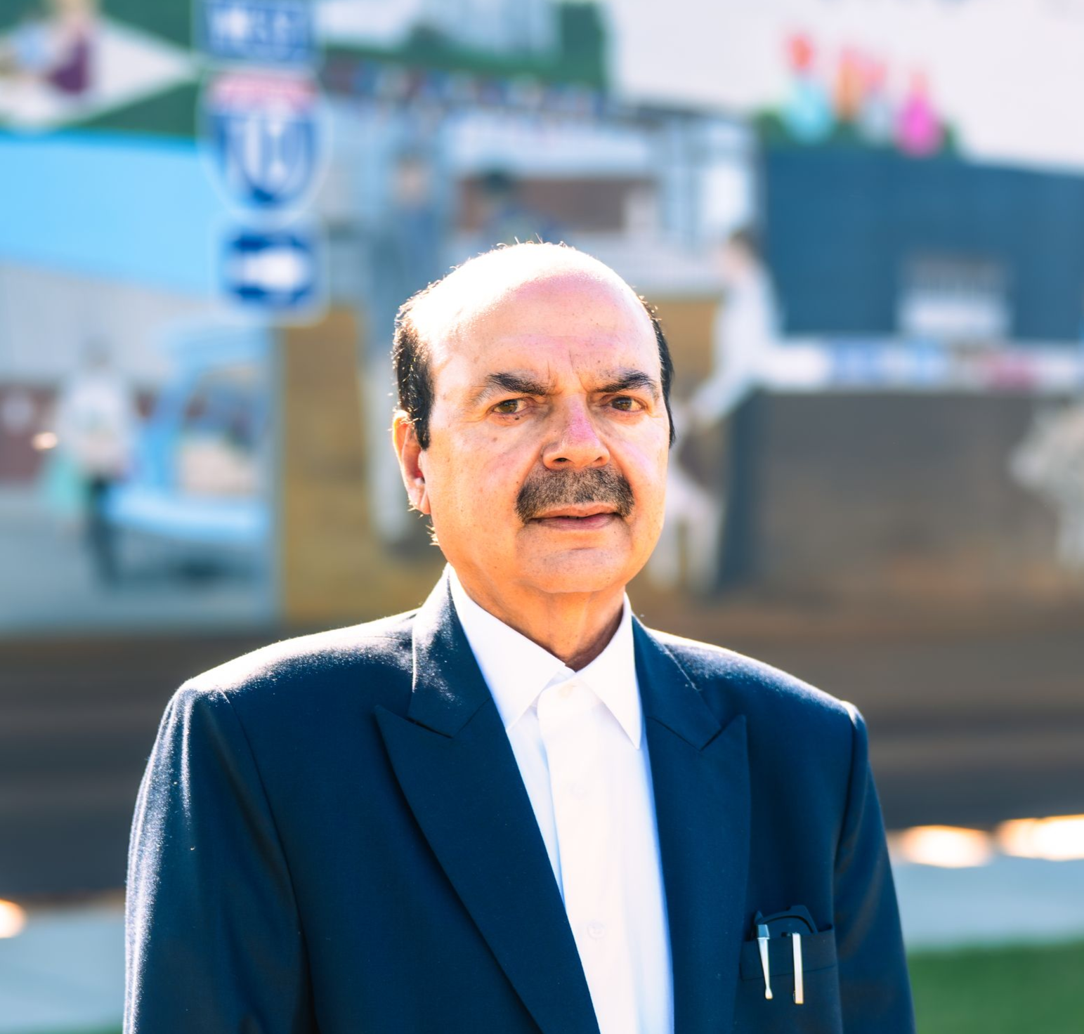 Man in a dark suit and white shirt, standing outside with a mural background.