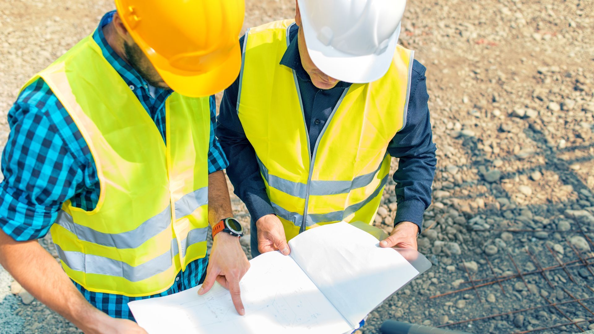 Deux ouvriers du bâtiment, vêtus de gilets haute visibilité et de casques de chantier, examinent ensemble des plans sur un site gravillonné.