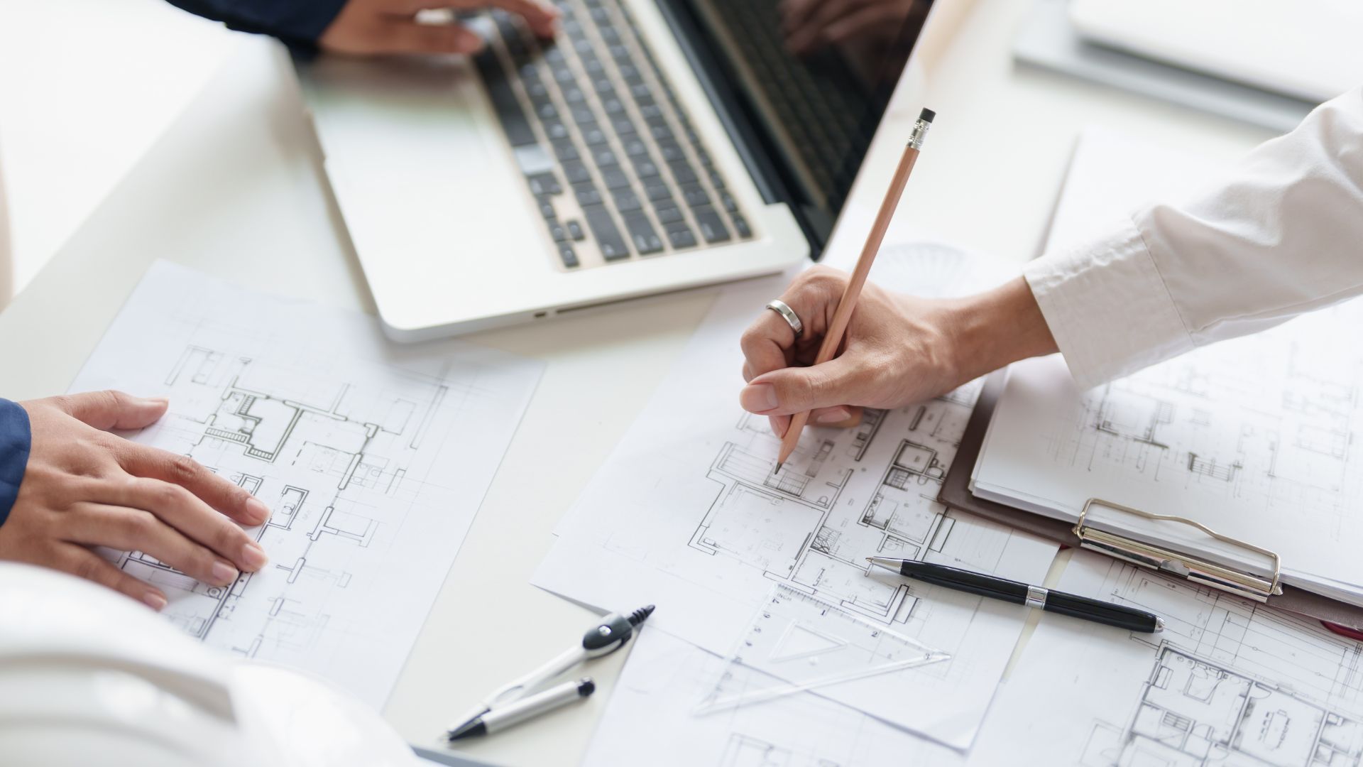 Deux personnes collaborent à des plans architecturaux à un bureau, équipées d'un ordinateur portable et d'outils de dessin.