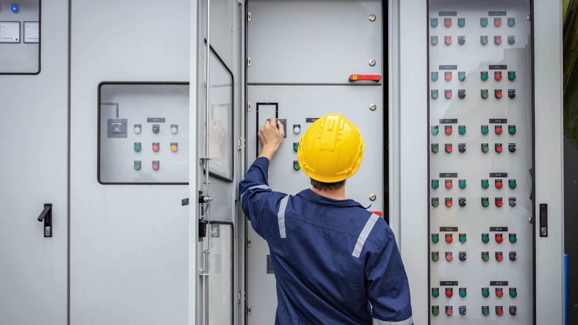 Un ouvrier portant un casque jaune et un uniforme bleu actionne un grand panneau de commande électrique industriel.